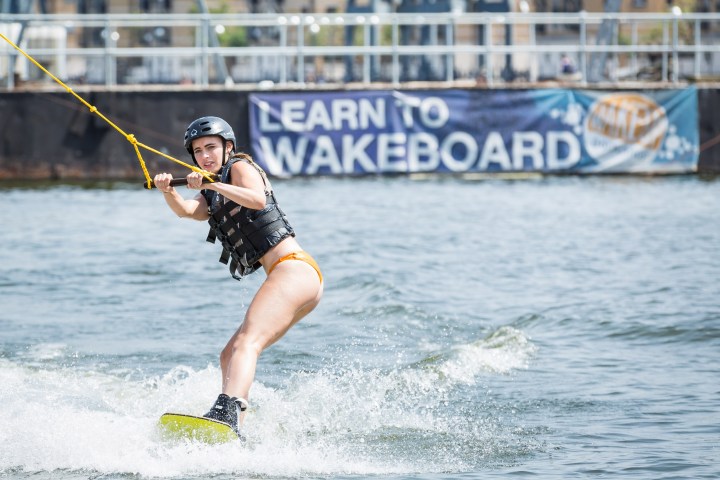 Person wakeboarding on water with helmet and life jacket, banner in background.