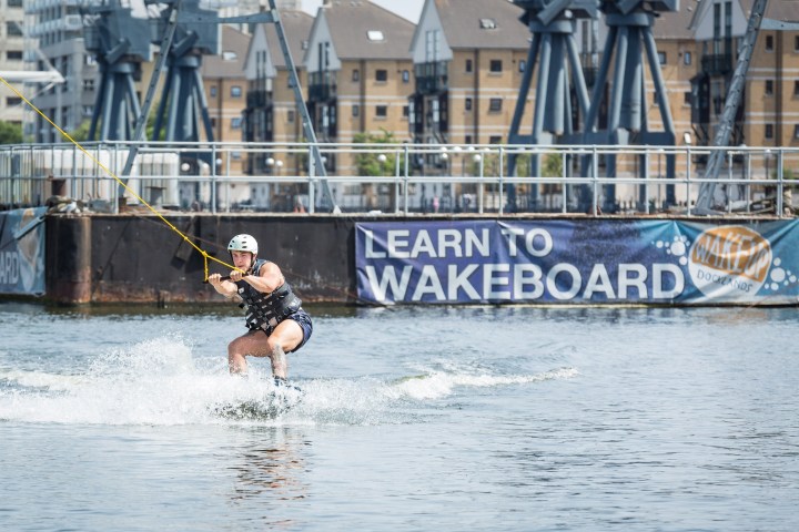 Person wakeboarding on water near docks with a 'Learn to Wakeboard' sign.