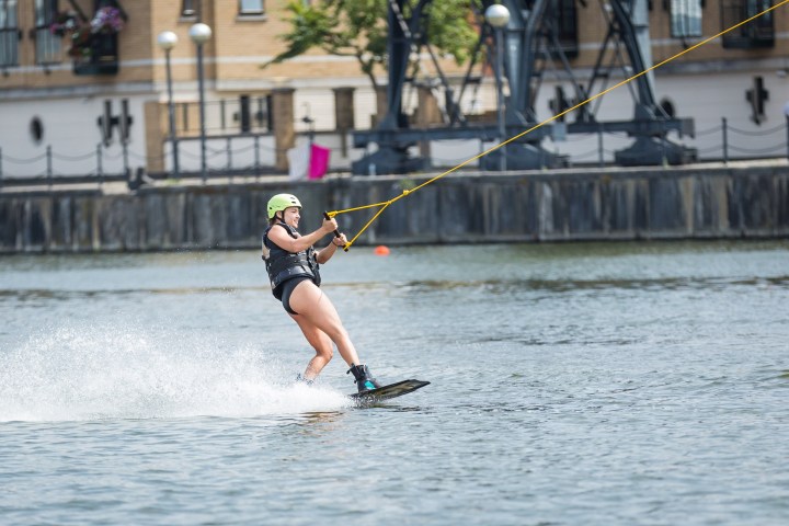 Person wakeboarding on a river with a yellow helmet and life vest, buildings in the background.