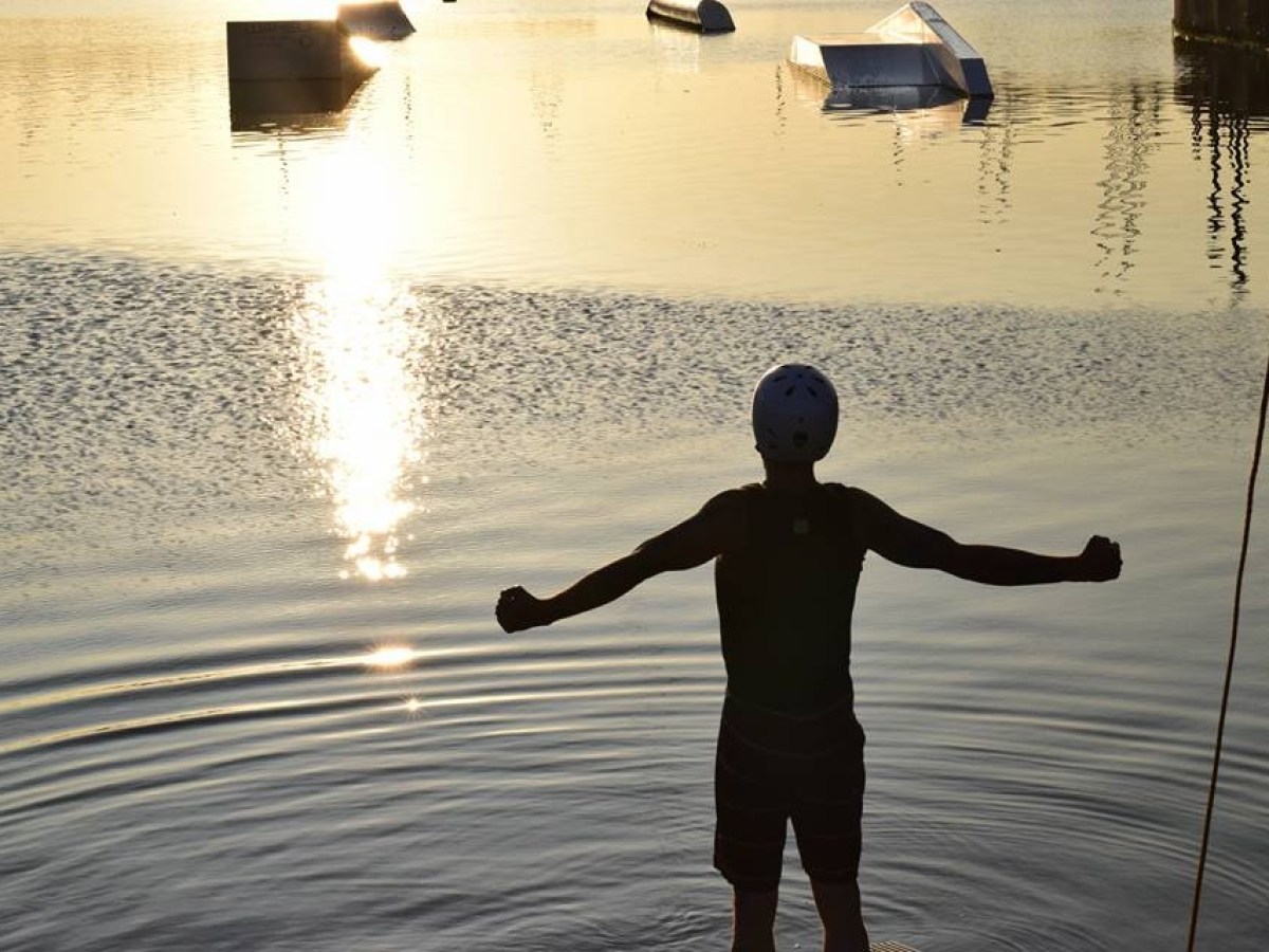 Silhouette of a person raising arms by a lake with obstacles at sunset.