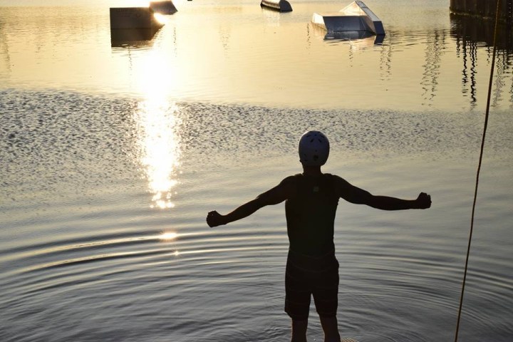 Silhouette of a person raising arms by a lake with obstacles at sunset.