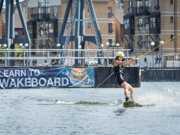 Person wakeboarding on water, wearing helmet and life jacket, with urban buildings in the background.