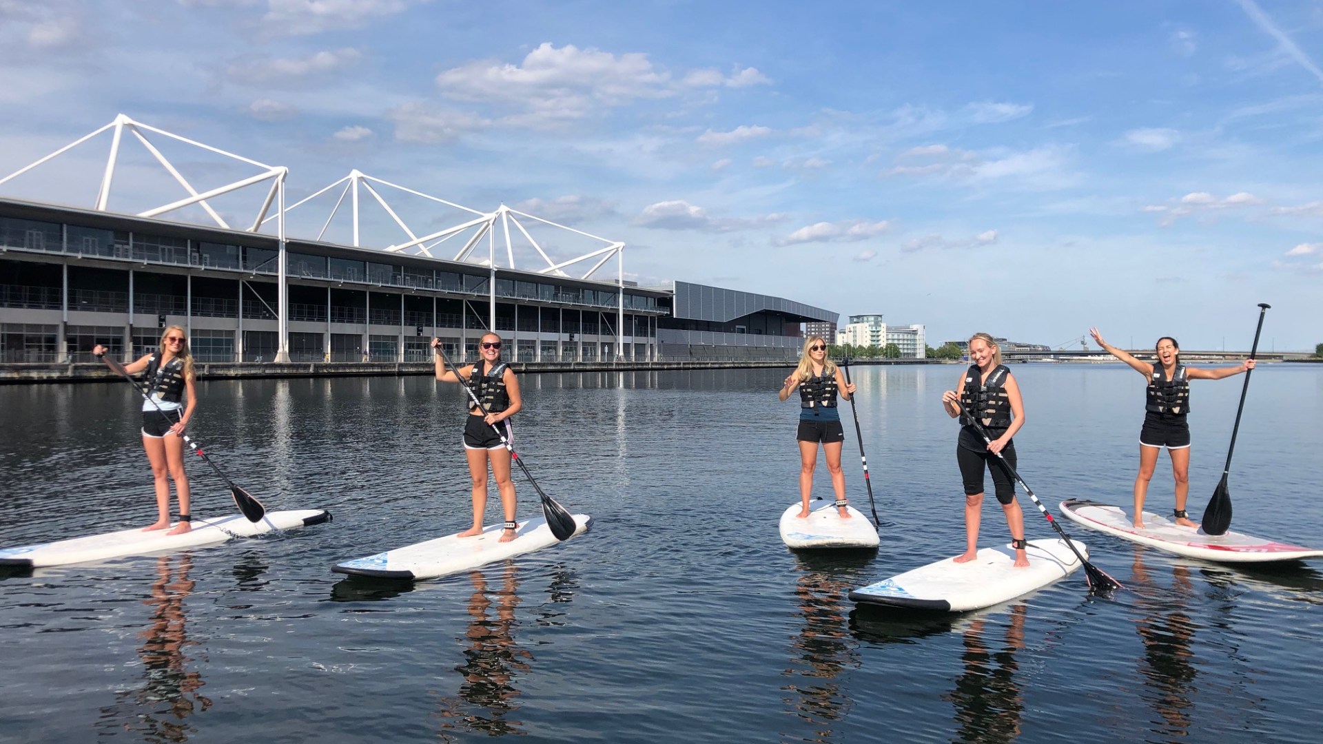 Five people paddleboarding on calm water near a modern building.