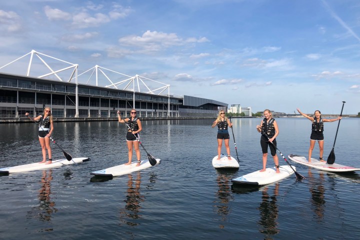 Five people paddleboarding on calm water near a modern building.