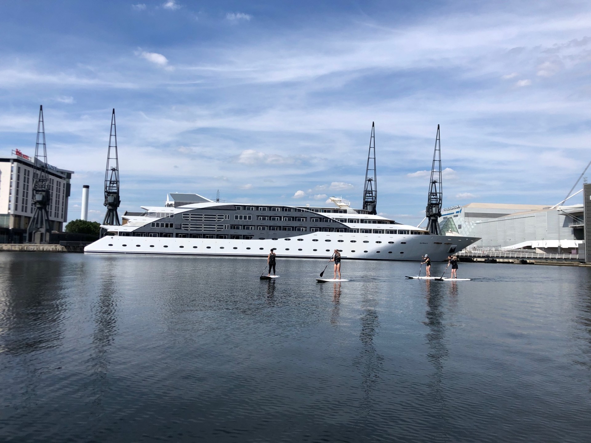 People paddleboarding near a large white yacht docked by cranes.