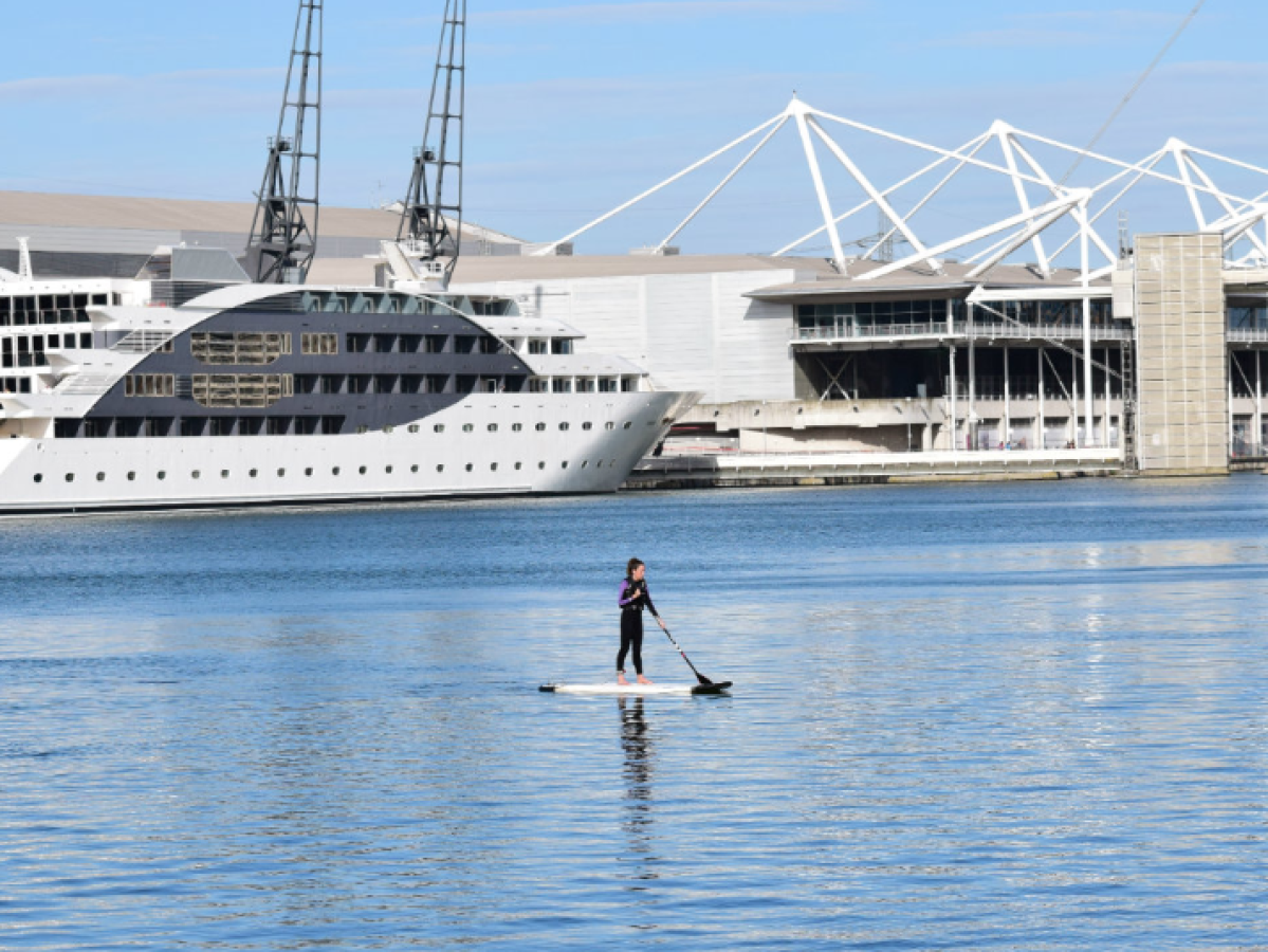 Person paddleboarding on water near large ship and white building.