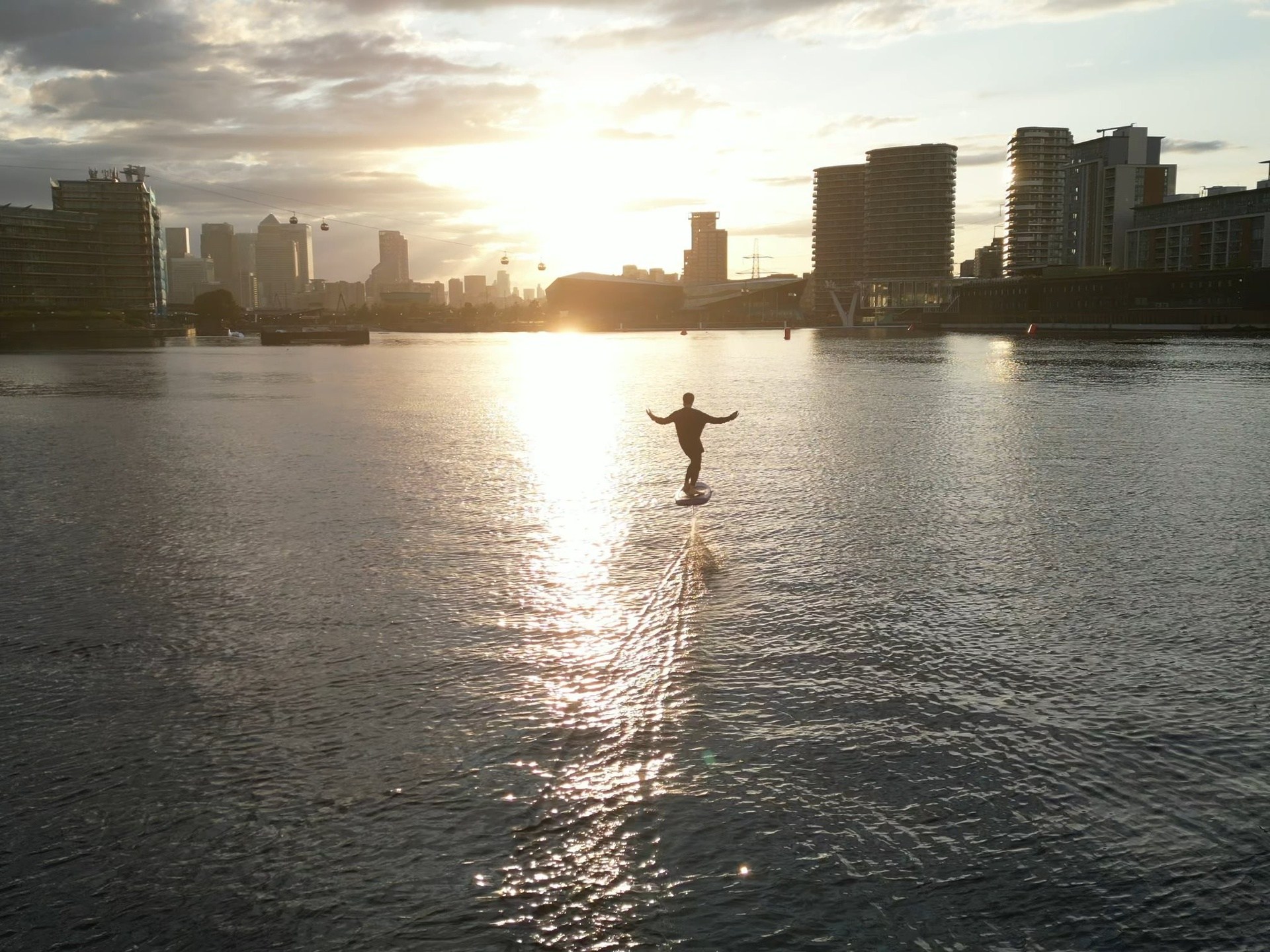 Person rides hoverboard on water at sunset, city skyline in background.