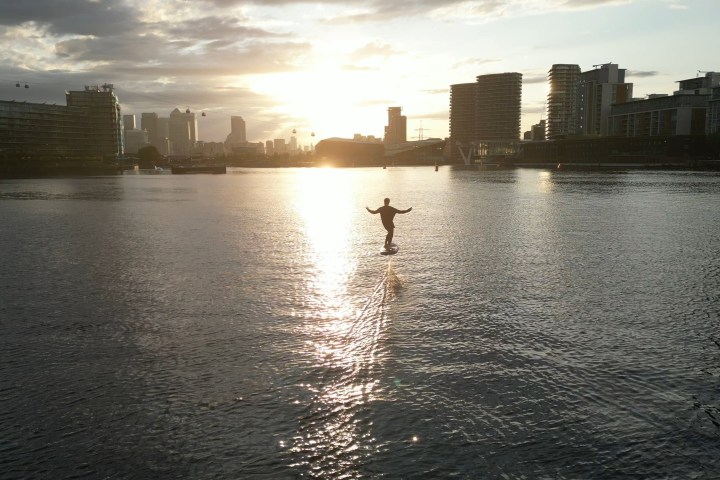 Person rides hoverboard on water at sunset, city skyline in background.