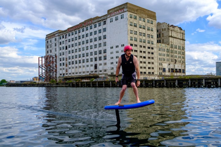 Person in a pink helmet rides an electric hydrofoil skateboard on a river near an old industrial building.