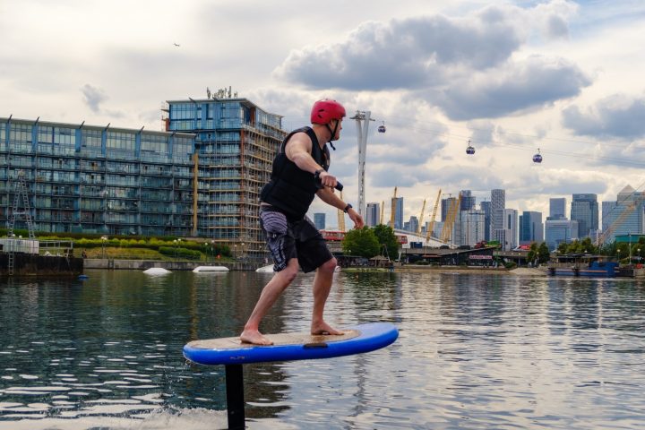 Person riding an electric hydrofoil board on a river with cityscape and cable cars in the background.