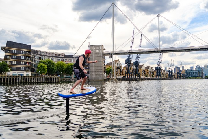 Person rides an electric hydrofoil board on a river near industrial cranes and a bridge.
