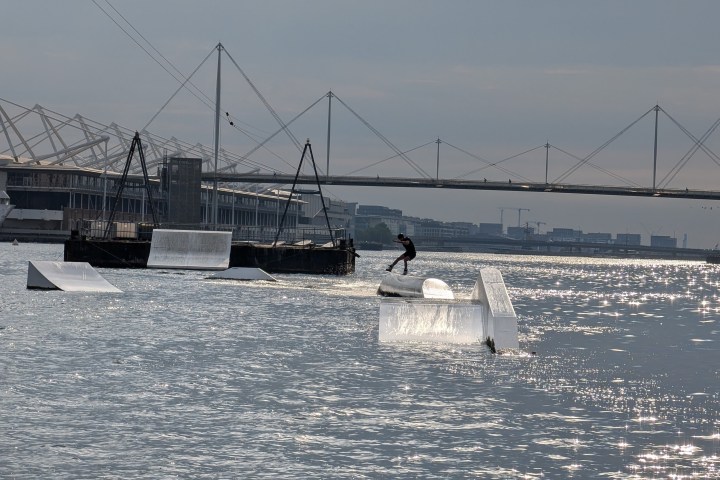 Person wakeboarding on a river with ramps and a bridge in the background.