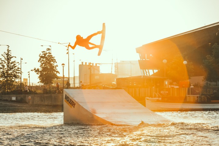 Wakeboarder performing a jump at sunset over a ramp on water.
