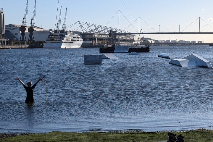 Person wades in river with arms up, near floating structures, bridge and cityscape in background.