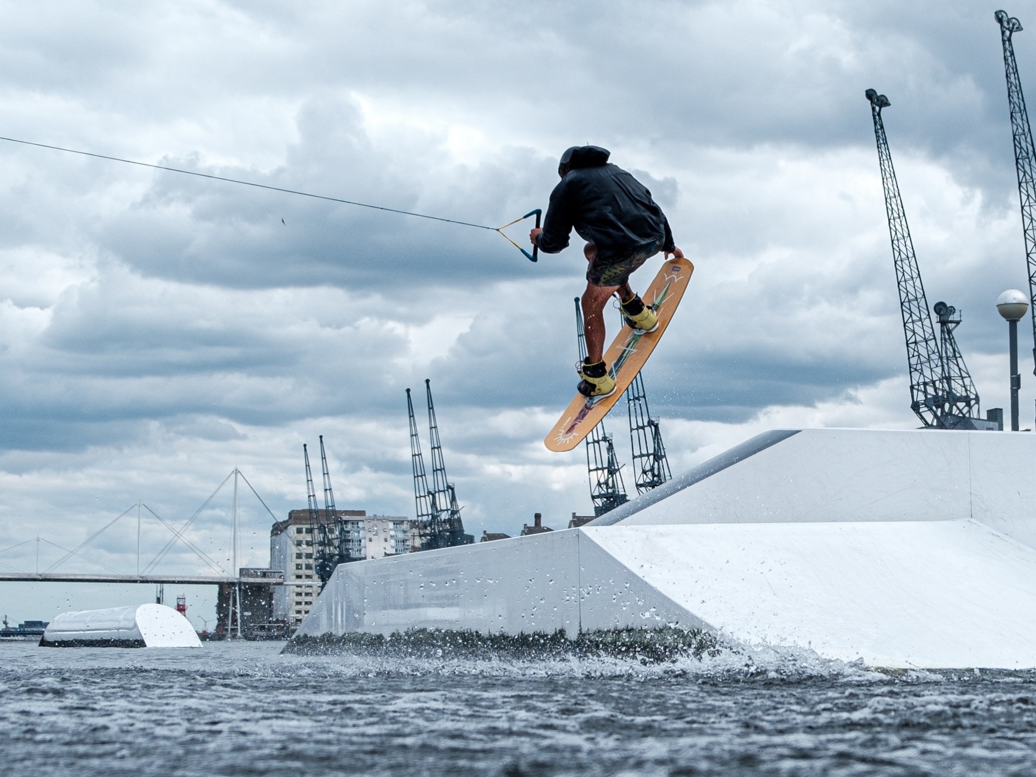 Person wakeboarding, mid-air trick, cloudy sky, cranes in background.