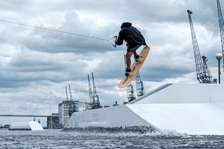 Person wakeboarding, mid-air trick, cloudy sky, cranes in background.