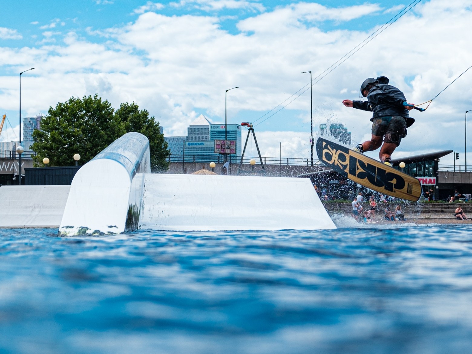 Person wakeboarding on water with a rail and city cable cars in the background.