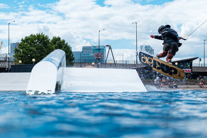 Person wakeboarding on water with a rail and city cable cars in the background.