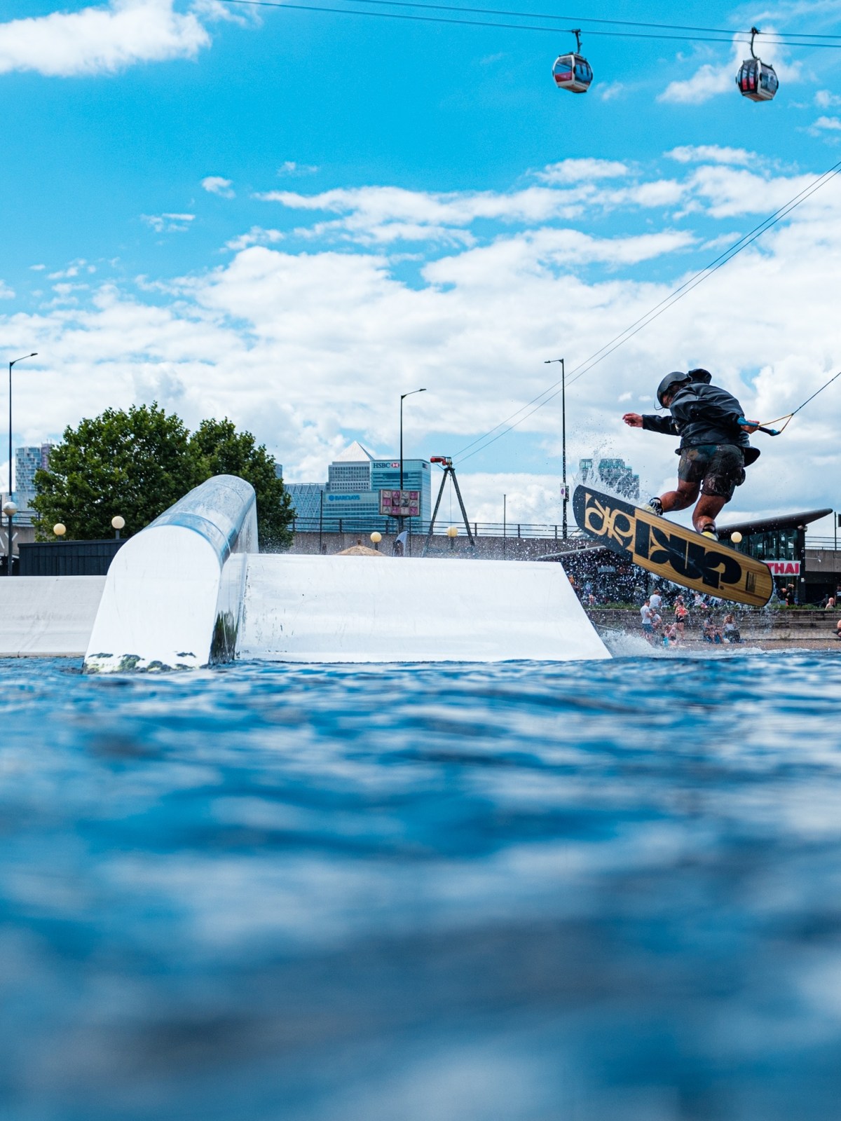 Person wakeboarding on water with a rail and city cable cars in the background.