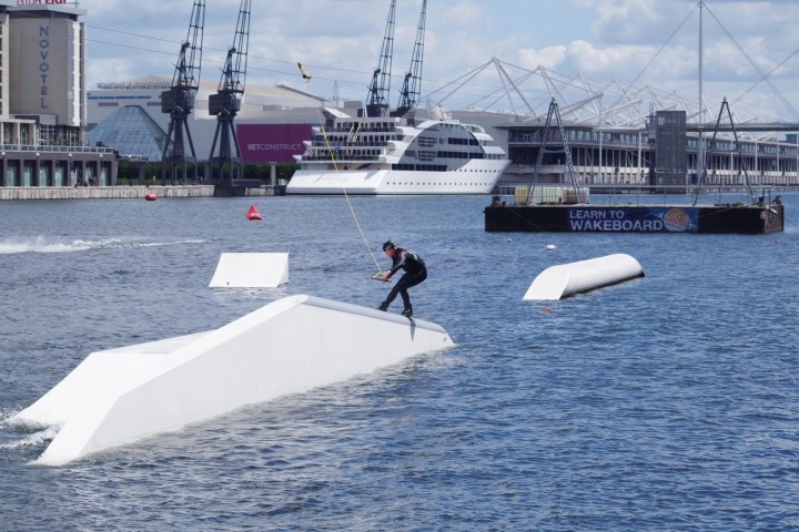 Person wakeboarding on water with ramps and buildings in background.