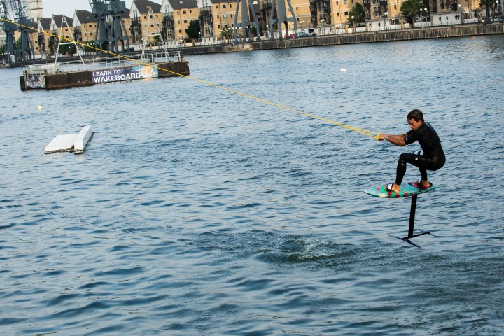Person wakeboarding on water with a hydrofoil, buildings and 'Learn to Wakeboard' sign in background.