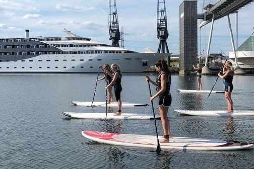 Five people paddleboarding on calm water near a large yacht and cranes.