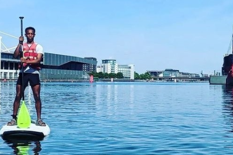 Person paddleboarding on a calm river with urban buildings and a red ship in the background.