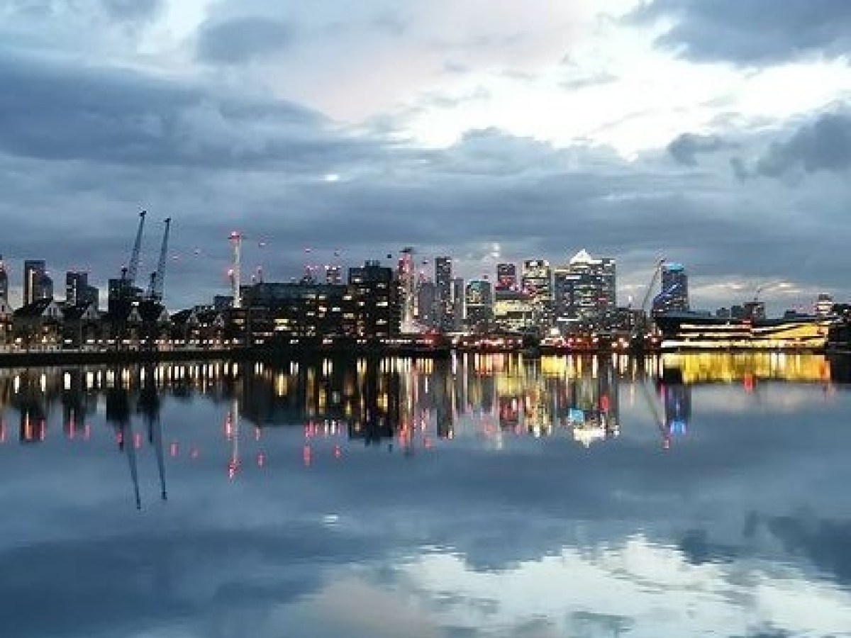 City skyline at dusk reflected in calm water with cranes and a docked ship.