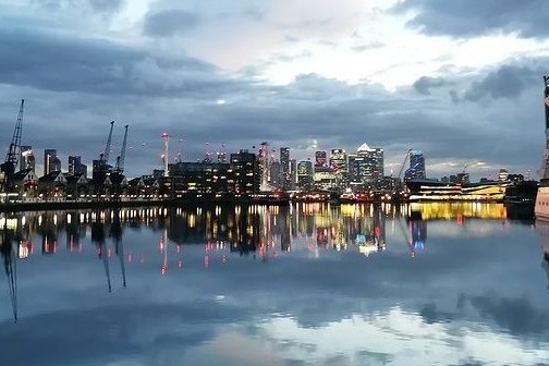 City skyline at dusk reflected in calm water with cranes and a docked ship.