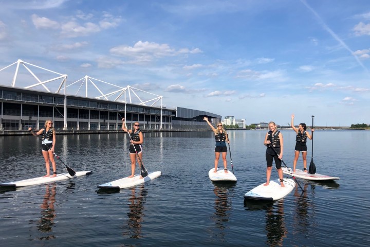 Five people paddleboarding on a calm river near modern buildings.