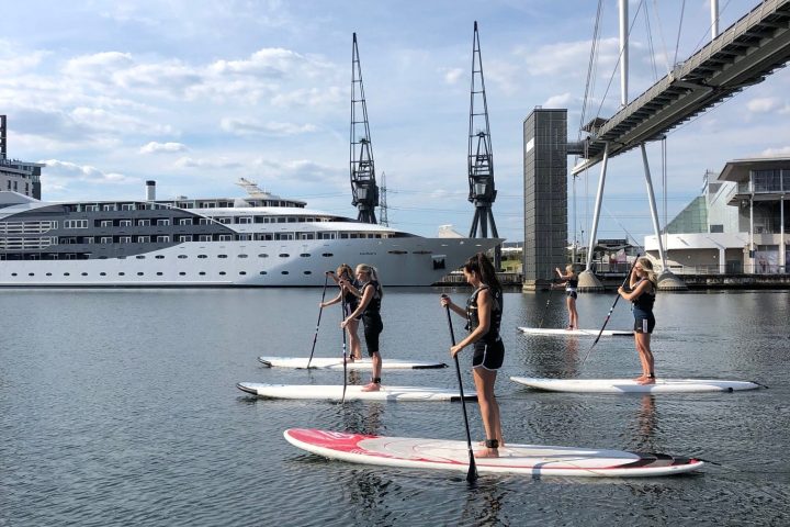 People paddleboarding on a river near a large ship and bridge.
