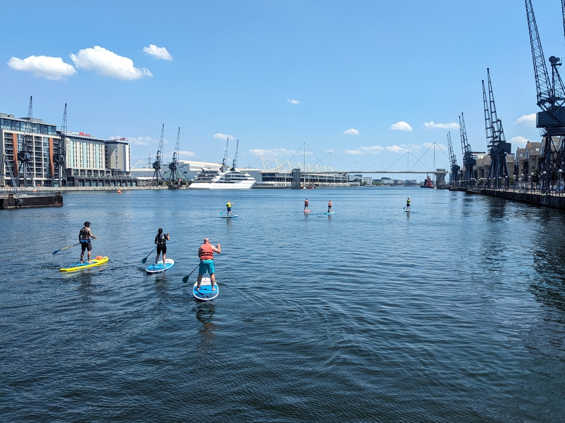 People paddleboarding on a calm urban river with cranes in the background.