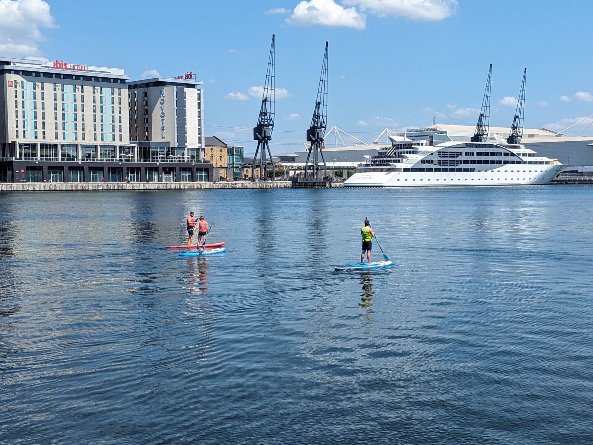 Two people paddleboarding on calm water near a large yacht and buildings.