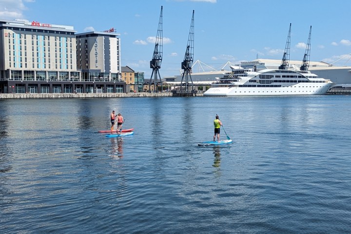 Two people paddleboarding on calm water near a large yacht and buildings.