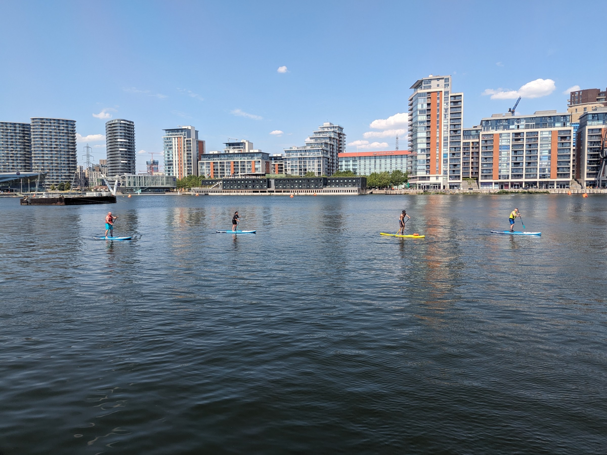 Four people paddleboarding on a calm river with city buildings in the background.